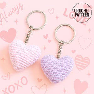 Peut inclure: Deux porte-cl&eacute;s en forme de c&oelig;ur au crochet, un rose clair et un lavande, avec des anneaux et des cha&icirc;nes argent&eacute;s. Le fond est rose avec des c&oelig;urs, des &eacute;toiles et les mots "Always" et "XOXO". Un cercle indique "CROCHET PATTERN".
