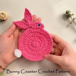 May include: A handmade pink crochet bunny coaster with blue eyes and a white tail. The coaster is held in two hands. The text "Bunny Coaster Crochet Pattern" is at the bottom of the image.