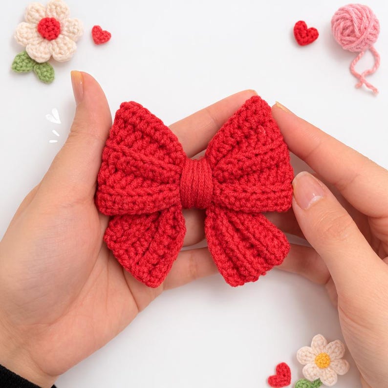 May include: A bright red crochet bow featuring a textured, ribbed stitch pattern with a central knotted yarn detail. The bow is constructed from yarn and displays a symmetrical shape with two large loops and two smaller tails extending downwards.