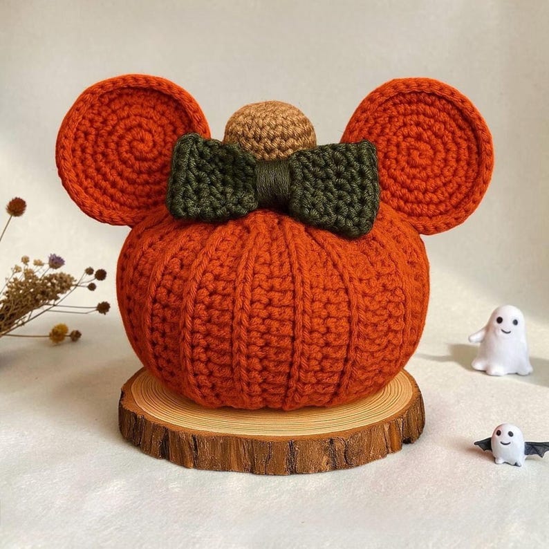 May include: A crocheted orange pumpkin with two large round ears, a dark green bow, and a small tan sphere on top. The pumpkin sits on a circular wooden slice. Two small white ghost figurines are placed nearby on the surface.