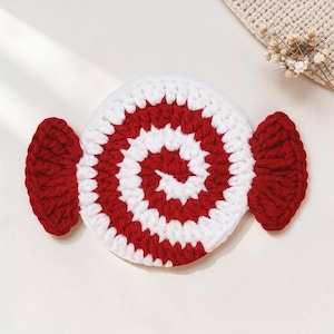 May include: A handmade crochet coaster in the shape of a candy. The coaster features a red and white spiral design in the center, with red candy wrapper ends. The coaster is made of soft yarn and is perfect for protecting surfaces from hot or cold drinks.