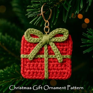 Puede incluir: Adorno de regalo de Navidad hecho a mano de ganchillo rojo con lazo y cintas verdes. El adorno está suspendido por un anillo dorado y se encuentra sobre un fondo de agujas de pino verdes. El texto "Christmas Gift Ornament Pattern" está en la parte inferior.