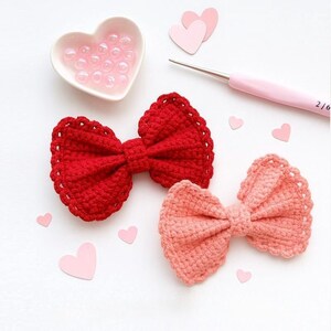 Pu&ograve; includere: Due fiocchi realizzati all'uncinetto a mano, uno rosso vivace e uno color pesca tenue, con un design a bordo smerlato. Un piattino a forma di cuore contiene piccole perline traslucide e un uncinetto rosa &egrave; posizionato vicino a cuori di carta.