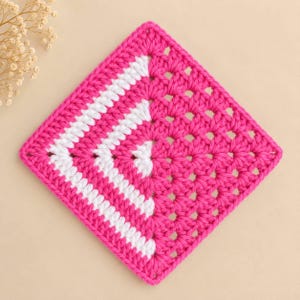 May include: A square crochet granny square featuring a vibrant hot pink yarn base with a contrasting white chevron pattern running diagonally across the left side. The piece is constructed with a classic cluster stitch technique throughout the design.