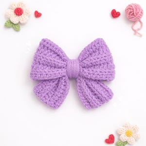 May include: A lavender crochet bow with a central knotted band. The bow features a textured, ribbed stitch pattern. Surrounding the bow are small decorative elements including two crocheted flowers with white petals and a pink yarn ball.