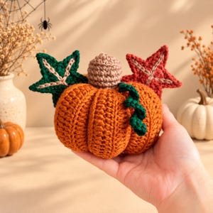 Puede incluir: Una calabaza naranja tejida a ganchillo con un tallo marrón claro, un zarcillo verde oscuro y dos hojas decorativas en forma de estrella en verde oscuro y rojo oscuro. La calabaza tiene una textura acanalada y se sostiene en la palma.