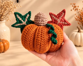 May include: A crocheted orange pumpkin featuring a light brown stem, a dark green curly vine, and two decorative star-shaped leaves in dark green and dark red. The pumpkin has a ribbed texture created by yarn stitches and is held in a palm.