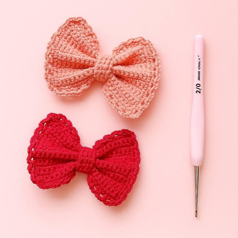 Pu&ograve; includere: Due fiocchi realizzati a mano all'uncinetto, uno rosa chiaro e uno rosso brillante, posizionati accanto a un uncinetto rosa chiaro con la scritta 2/0 stampata sul manico. Gli oggetti sono disposti su una superficie rosa chiaro.