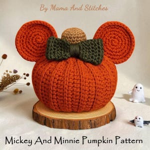 May include: A crocheted pumpkin in orange with circular ears and a green bow, resembling Mickey Mouse. The pumpkin sits on a wooden slice, with the text "Mickey And Minnie Pumpkin Pattern" at the bottom. The text "By Mama And Stitches" is at the top.