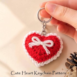 May include: A red heart-shaped crochet keychain featuring a white crocheted border and a small white yarn bow attached to the center. The heart is connected to a silver metal split ring by a short chain. The text "Cute Heart Keychain Pattern" is visible.