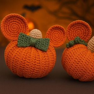 May include: Two orange crocheted pumpkins with round ears, one featuring a green bow and a small tan stem, and the other with a green stem and tan accent. The pumpkins have a ribbed texture and are crafted from yarn in a rounded, plush shape.