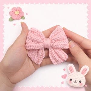 May include: A light pink crocheted bow with a textured ribbed pattern, accompanied by a small crocheted pink flower with a yellow center and a small white crocheted rabbit head with pink ears and black eyes, all featuring a soft yarn construction.
