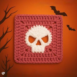May include: A square crochet granny square featuring a white skull motif centered on a dusty rose pink background. The skull design is created with white yarn stitches, contrasting against the textured pink yarn of the square border.