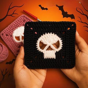 May include: A square crochet motif featuring a white skull design centered on a black background. A second matching square motif in dusty rose with a white skull is visible nearby, alongside a red crochet hook and a ball of matching yarn.