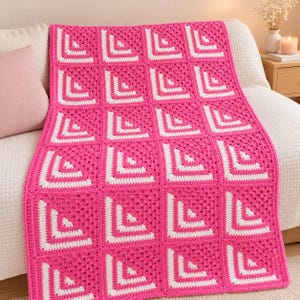 May include: A rectangular crochet blanket featuring a repeating pattern of square motifs. Each square is composed of alternating rows of vibrant hot pink and crisp white yarn, arranged to create a geometric corner design across the entire piece.