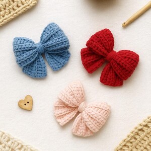 May include: Three handmade crochet bows in blue, red, and light pink colors. Each bow features a textured, ribbed yarn construction with a central knot detail. A small, light brown wooden heart-shaped button with two holes is positioned nearby.