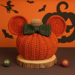 May include: A crocheted orange pumpkin with two round orange ears, a small tan sphere on top, and a dark green bow. The pumpkin sits on a circular wooden slice, flanked by a small green sphere on the left and a small red sphere on the right.