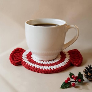 May include: A white ceramic mug filled with dark liquid sits on a red and white crocheted coaster. The coaster has a candy cane design. A sprig of holly with red berries and a pinecone are in the foreground.