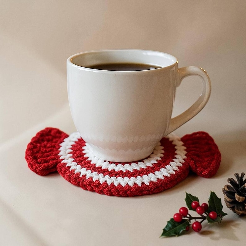May include: A white ceramic mug filled with dark liquid sits on a red and white crocheted coaster. The coaster has a candy cane design. A sprig of holly with red berries and a pinecone are in the foreground.