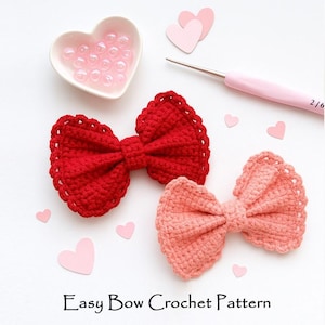May include: Two crocheted bow hair accessories, one red and one peach, with scalloped edges, are displayed on a white surface. Pink heart-shaped cutouts and a heart-shaped dish with pink beads are scattered around. The text "Easy Bow Crochet Pattern" is at the bottom.