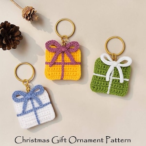 May include: Three crocheted gift-shaped keychains in yellow, white, and green, each with a contrasting bow and gold-colored ring. The keychains are decorated with white or purple yarn. The text "Christmas Gift Ornament Pattern" is at the bottom.