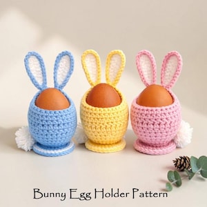 Op de afbeelding: Drie gehaakte konijnen eierhouders in blauw, geel en roze, elk met een bruin ei. De houders hebben konijnenoren en een pluizige staart. De tekst "Bunny Egg Holder Pattern" staat onderaan.
