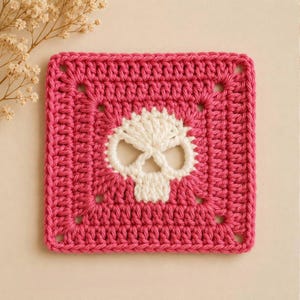 May include: A square crochet granny square featuring a vibrant hot pink border and a central white skull motif. The piece is crafted with visible yarn stitches, creating a textured surface with a distinct geometric pattern and a clear skull design.