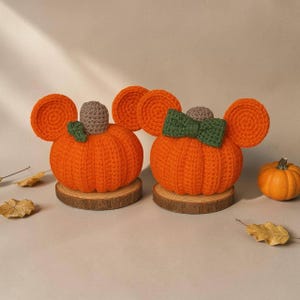 May include: Two crocheted orange pumpkins with round ears, each resting on a circular wooden base. One pumpkin features a small green leaf and a brown stem, while the other has a green bow and a brown stem. A small real pumpkin sits nearby.