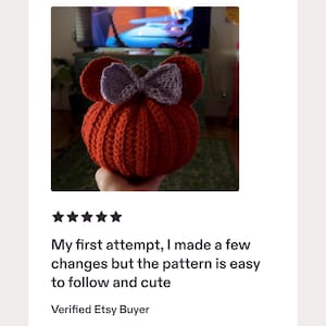 May include: A round, orange, crocheted pumpkin-shaped object featuring two rounded ears on top and a light purple crocheted bow centered between them. The pumpkin has a textured, ribbed surface and a small brown stem visible at the very top center.