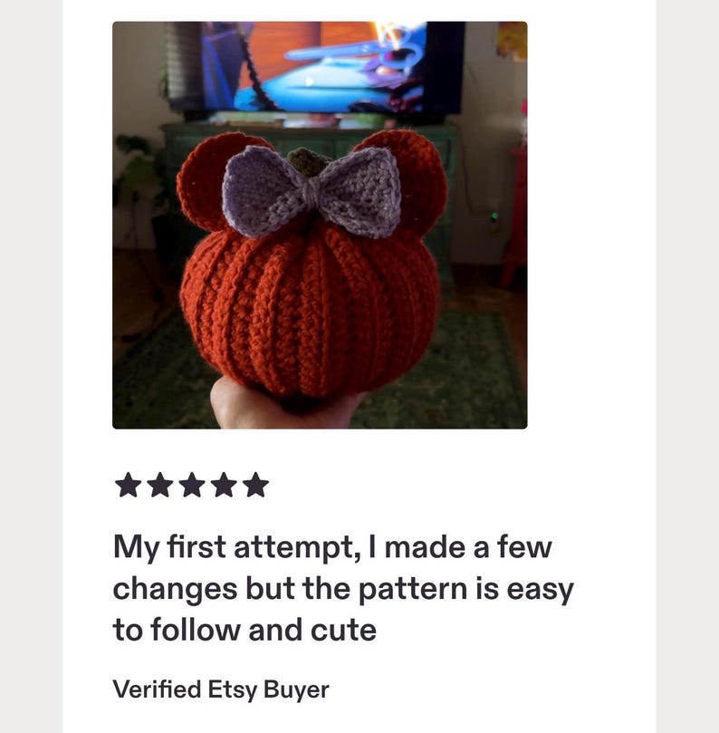 May include: A round, orange, crocheted pumpkin-shaped object featuring two rounded ears on top and a light purple crocheted bow centered between them. The pumpkin has a textured, ribbed surface and a small brown stem visible at the very top center.