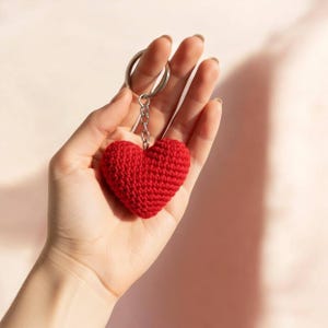 Peut inclure: Un porte-cl&eacute;s en forme de c&oelig;ur rouge crochet&eacute;. Le charm en forme de c&oelig;ur est attach&eacute; &agrave; un porte-cl&eacute;s et une cha&icirc;ne argent&eacute;s. Le porte-cl&eacute;s est tenu dans une main sur un fond rose clair.
