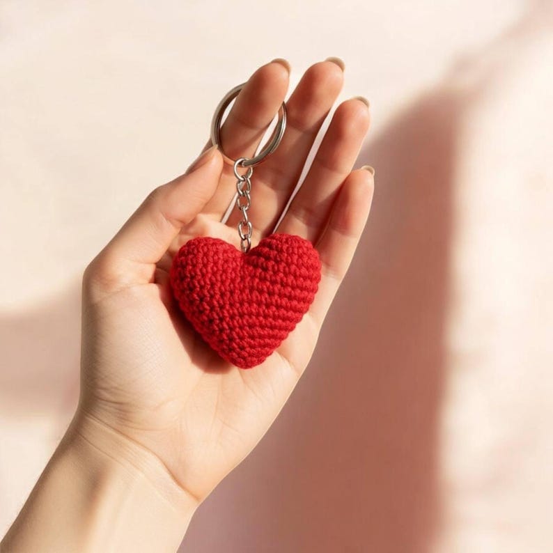 Peut inclure: Un porte-cl&eacute;s en forme de c&oelig;ur rouge crochet&eacute;. Le charm en forme de c&oelig;ur est attach&eacute; &agrave; un porte-cl&eacute;s et une cha&icirc;ne argent&eacute;s. Le porte-cl&eacute;s est tenu dans une main sur un fond rose clair.