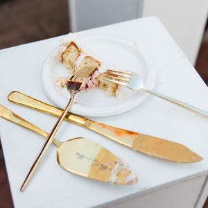 May include: A gold-toned cake server and two matching forks resting on a white plate containing a slice of cake with white frosting and pink filling. The cake server features an engraved script text on its surface, and the utensils are arranged in a cross pattern on the plate.