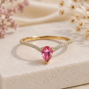 May include: A gold ring featuring a pear-shaped pink gemstone at the center of a V-shaped band. The band is set with a row of small, clear, round-cut stones that extend from the central pink stone along both sides of the thin, polished gold shank.