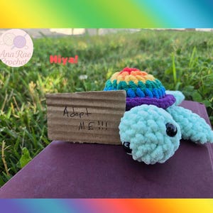 Op de afbeelding: Een gehaakte schildpad met een regenboogkleurig schild en lichtblauw lichaam. Een klein kartonnen bordje leest "Adopt ME!!!". De schildpad zit op een paars oppervlak met gras op de achtergrond.