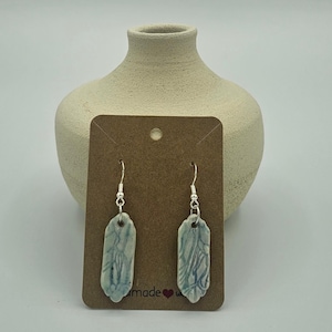 May include: Handmade ceramic earrings with a light blue and white design, hanging from silver-colored hooks. The earrings are displayed on a brown card with the word "made" and a heart symbol. A beige vase is in the background.