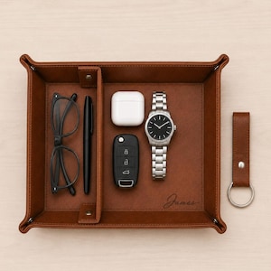 May include: A rectangular brown valet tray with a divider, featuring the name James embossed on the surface. The tray contains black spectacles, a black pen, white wireless earbuds, a black car key fob, and a silver wristwatch. A matching brown leather keyring is placed next to the tray.