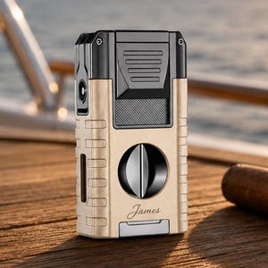 May include: A rectangular cigar lighter and cutter combination tool featuring a matte gold finish with black accents. The device includes a circular blade mechanism, a textured grip, and the name James engraved in a cursive script on the front.