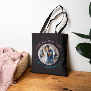 May include: Black tote bag featuring a disco ball design with a person inside. The text "KISS ALL THE TIME. DISCO, OCCASIONALLY." is printed in pink and blue. The bag has black handles.