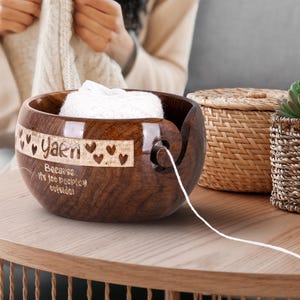 Könnte beinhalten: Eine dunkelbraune Garnschale aus Holz mit dem Wort "Yarn" und Herzmotiven auf der Seite. Die Schale enthält weiße Wolle, wobei ein Faden durch eine Spirale verläuft. Der Text "Because it's too peopley outside!" ist ebenfalls eingraviert.