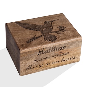 Op de afbeelding: Houten kist met een kolibrie-gravering op het deksel. De kist is gegraveerd met de naam "Matthew", data en de zin "Always in our hearts". Het hout heeft een natuurlijke nerf en een warme bruine kleur.