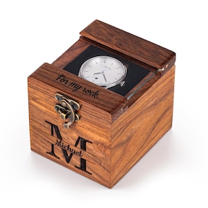 May include: A wooden watch box with a silver watch inside. The box is engraved with the name "Michael" and the phrase "For my rock." The watch has a white face and a black leather band. The box is closed with a metal clasp.