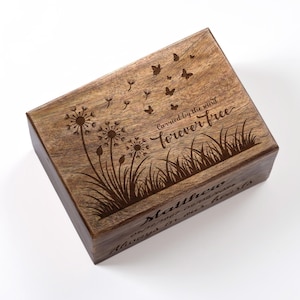 May include: A rectangular wooden box with engraved dandelions, butterflies, and grass. The text "Carried by the wind forever free" is above the grass. The box also has the name "Matthew" and dates engraved on it.