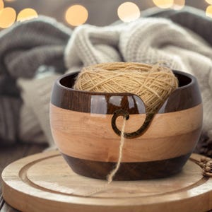 以下が含まれることがあります： 明るい茶色の糸玉が入った木製の糸入れ。ボウルは明るい木材と暗い木材の交互の帯でできています。湾曲したスロットから糸が出てきます。ボウルは木製のまな板の上に置かれています。