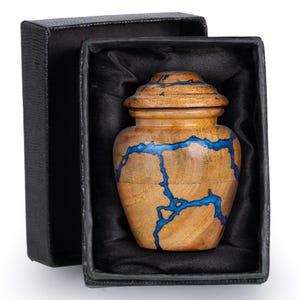 May include: A small wooden urn with a lid, presented in a black box. The urn is light brown with striking blue resin accents. The box is lined with black satin.