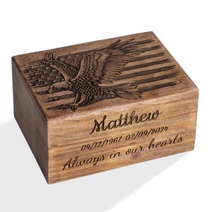 Può includere: Una scatola di legno con un disegno inciso della bandiera americana e un'aquila. La scatola è personalizzata con il nome "Matthew", le date e la frase "Always in our hearts". Il legno è di colore marrone chiaro.