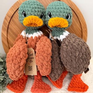 May include: Two handmade crochet duck plushies. Each duck has a green head, orange beak, and orange feet. One duck has brown wings, the other has orange wings. Both have a white and brown collar. The tag reads "Knot Alone Co."