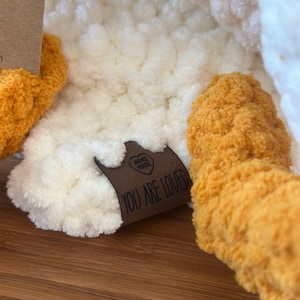 Peut inclure: Gros plan sur un jouet en peluche fait &agrave; la main, avec un corps cr&egrave;me et des pieds orange. Une &eacute;tiquette marron avec les mots "YOU ARE LOVED" est attach&eacute;e au jouet. Le jouet est fait d'une mati&egrave;re douce et textur&eacute;e.