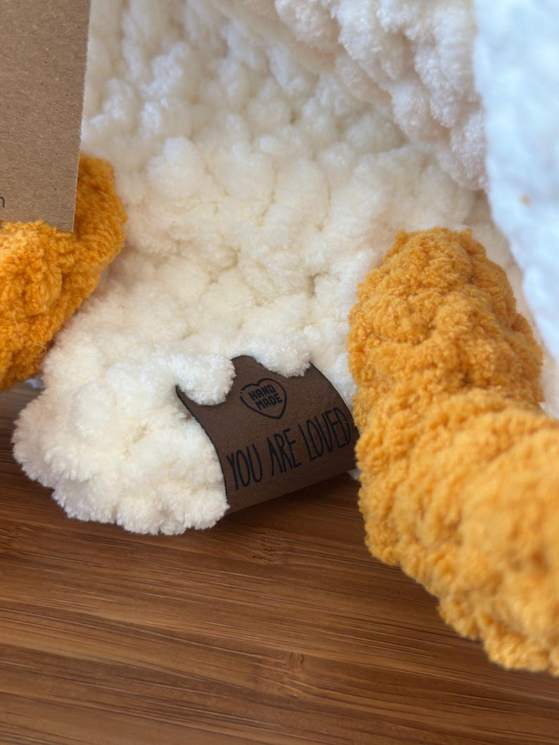 Peut inclure: Gros plan sur un jouet en peluche fait &agrave; la main, avec un corps cr&egrave;me et des pieds orange. Une &eacute;tiquette marron avec les mots "YOU ARE LOVED" est attach&eacute;e au jouet. Le jouet est fait d'une mati&egrave;re douce et textur&eacute;e.