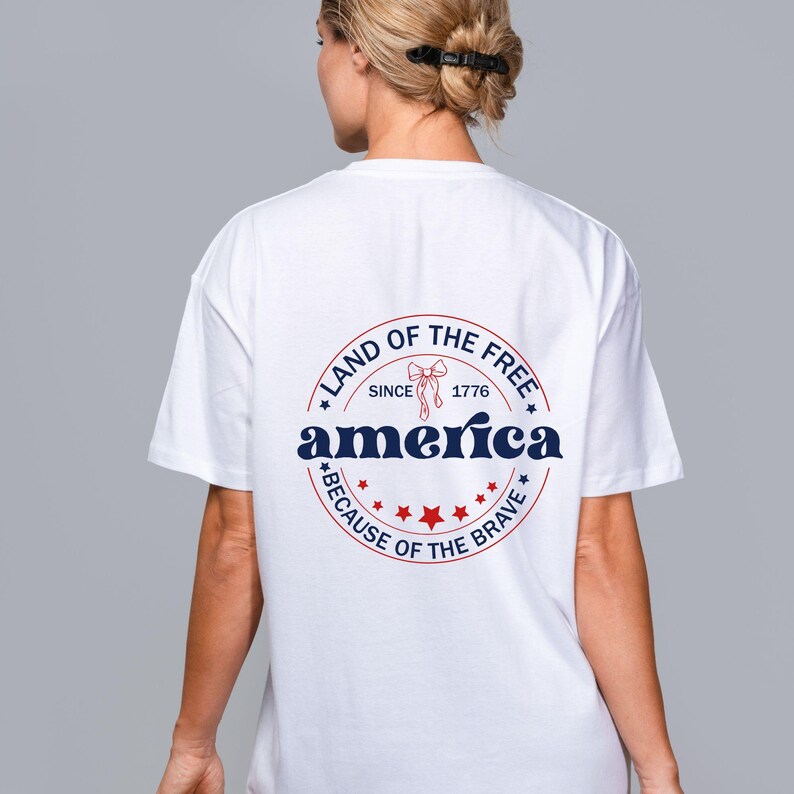 May include: White t-shirt with a circular graphic on the back. The graphic reads "Land of the Free" and "America" in navy blue, with red stars and a bow. The text "Because of the Brave" is also included.